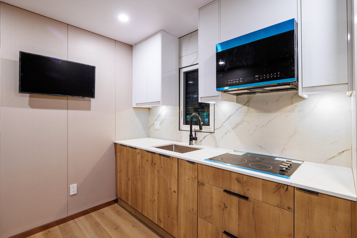 Compact exhibition kitchen display with a TV, pale wall panels and wood-tone cabinetry.