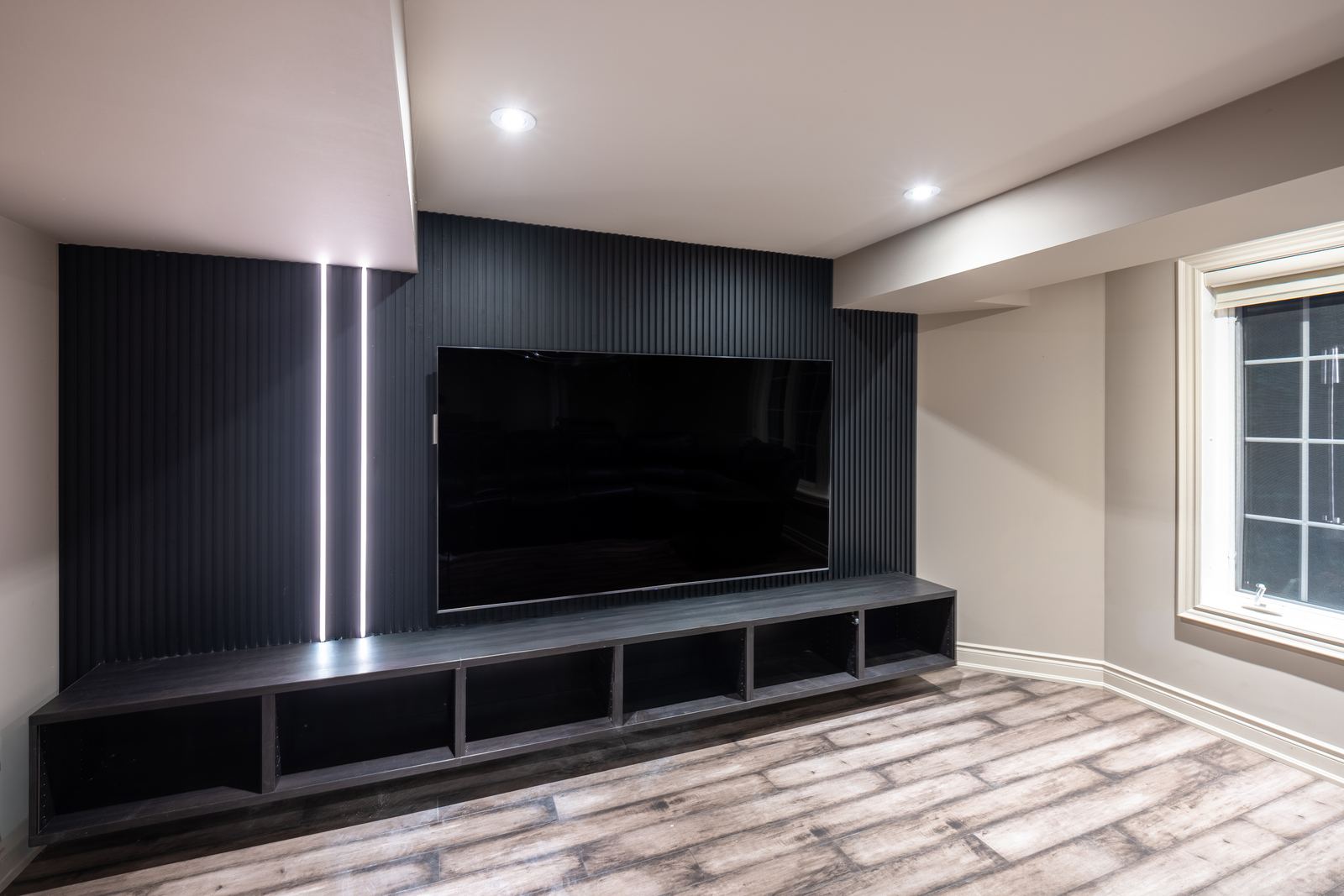 Angled view of matte black slat TV feature wall with vertical LED strips and long media console in Whitby Ontario by RichVaugh